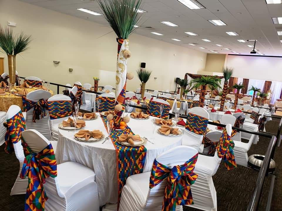 Grande salle de réception aux tables rondes, avec des chaises à ruban kente et des centres de table en herbes hautes