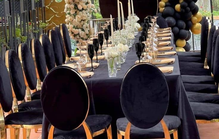 Longue table d’anniversaire à nappe noire, centres de table en fleurs blanches, chaises noires à cadre doré et guirlande de ballons noirs et dorés