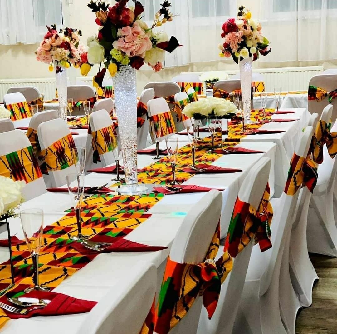 Longue table à chemin de table kente, hauts vases de fleurs et chaises blanches nouées de rubans kente