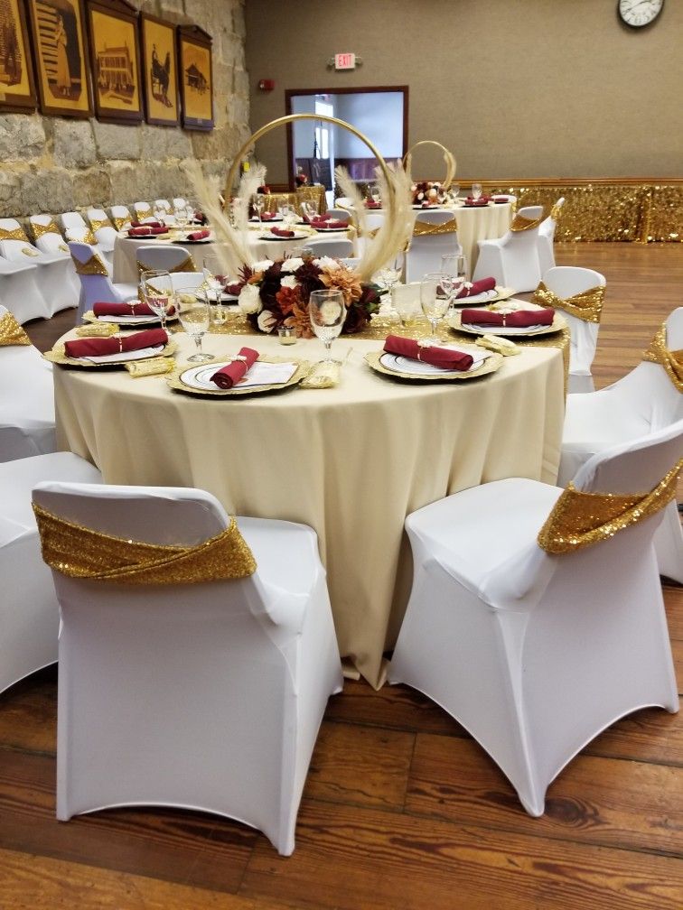 Table ronde à nappe crème, serviettes bordeaux et centre de table fleuri, chaises à housse blanche et ruban doré pailleté