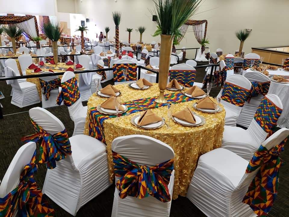 Table ronde à nappe dorée, chemin de table en tissu kente et chaises blanches nouées d’un ruban kente