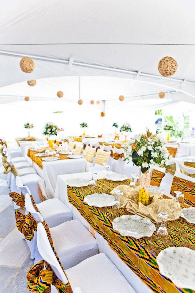 Intérieur d’une tente blanche aux longues tables ornées de chemins kente, de fleurs blanches et de boules en rotin suspendues