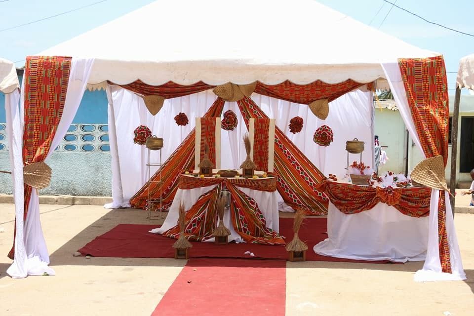 Tente blanche habillée de tissus kente rouge et orange, avec un tapis rouge et une table de cérémonie